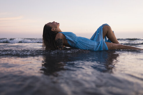 Junge tätowierte Frau in blauem Kleid, die bei Sonnenuntergang im Wasser am Meeresufer liegt - MAUF01723
