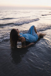 Young tattooed woman wearing blue dress lying in water at seashore by sunset - MAUF01724