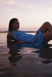 Young woman with tattoo wearing blue dress lying in water at seashore by sunset - MAUF01725