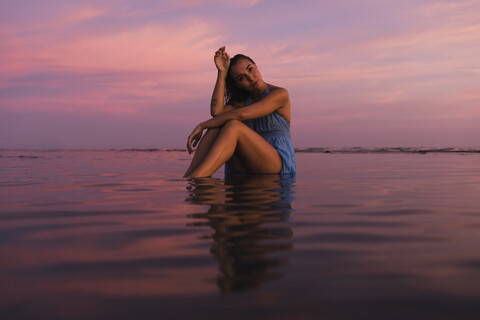 Profil einer jungen Frau, die bei Sonnenuntergang am Meer im Wasser sitzt, lizenzfreies Stockfoto