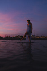 Young woman wearing summer dress standing in water at seashore by sunset splashing with water - MAUF01731