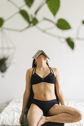 Young woman in lingerie sitting on chair at home covering her head with book - AFVF01676