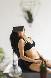 Young woman in lingerie sitting on chair at home covering her head with book - AFVF01677