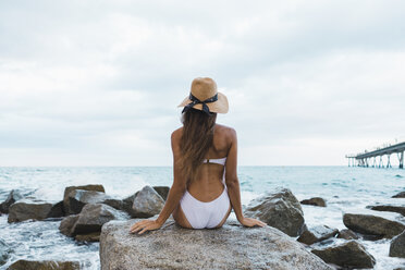 Rückansicht einer jungen Frau mit Badeanzug und Hut, die auf einem Felsen im Meer sitzt - AFVF01764