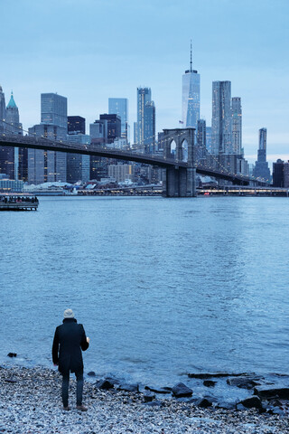 Mann blickt vom Flussufer aus auf die Brooklyn Bridge und die Skyline von Lower Manhattan, New York, USA, lizenzfreies Stockfoto