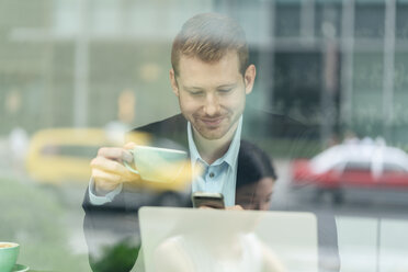 Geschäftsmann, der in einem Café sitzt, Kaffee trinkt und sein Smartphone benutzt, durch ein Fenster gesehen - ISF19726