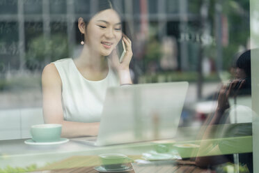 Businesswoman sitting in cafe, using smartphone and laptop, viewed through window - ISF19729