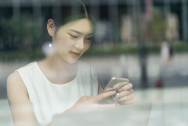 Businesswoman using smartphone and laptop, viewed through window - ISF19731