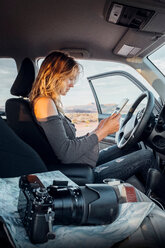 Junge Frau sitzt im Fahrzeug und schaut auf ein digitales Tablet, Spiegelreflexkamera auf dem Beifahrersitz, Mexican Hat, Utah, USA - ISF19733