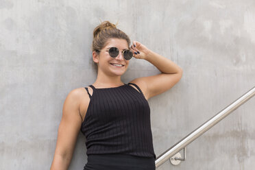 Porträt einer lächelnden jungen Frau mit Sonnenbrille, die an einer Betonwand lehnt - JUNF01509