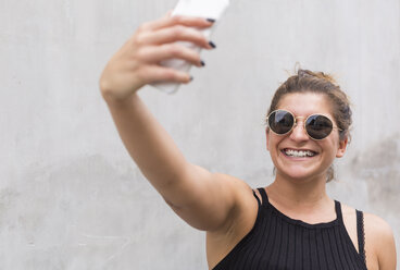 Porträt einer lachenden jungen Frau mit Sonnenbrille, die ein Selfie mit ihrem Smartphone macht - JUNF01513
