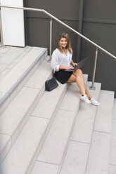 Porträt einer lächelnden Geschäftsfrau, die auf einer Treppe sitzt und einen Laptop benutzt - JUNF01524
