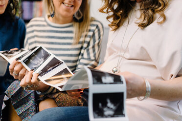 Pregnant woman and friends on sofa looking at ultrasound pictures, cropped - CUF46140