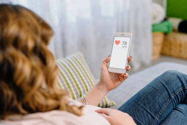 Pregnant woman on sofa looking at heart rate on smartphone, over shoulder view - CUF46148