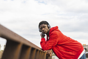 Young black man leaning on fence, listening music with headphones - AFVF01821