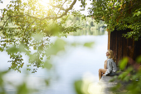 Entspannte Frau sitzt auf einem Holzsteg an einem abgelegenen See, lizenzfreies Stockfoto