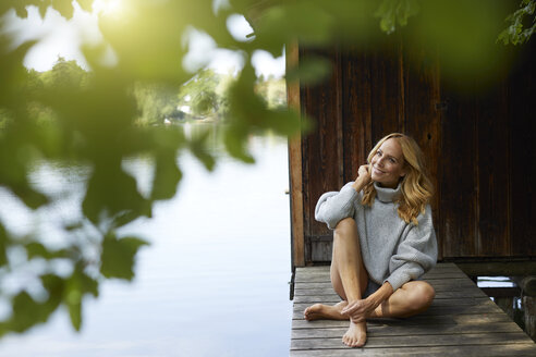 Lächelnde entspannte Frau, die auf einem Holzsteg an einem abgelegenen See sitzt - PNEF01047