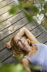Glückliche blonde Frau auf einem Holzsteg an einem See liegend - PNEF01066