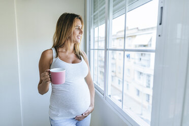 Pregnant woman looking out of window holding mug - KIJF02052