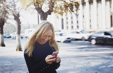 Lächelnde blonde Frau mit Smartphone in der Stadt - AZF00096