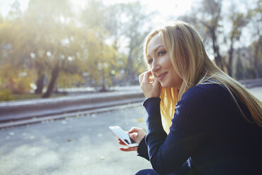 Portrait of content blond woman with smartphone in autumnal city park - AZF00098