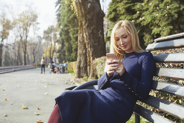 Porträt einer blonden Frau, die auf einer Bank im herbstlichen Stadtpark sitzt und ihr Smartphone benutzt - AZF00100