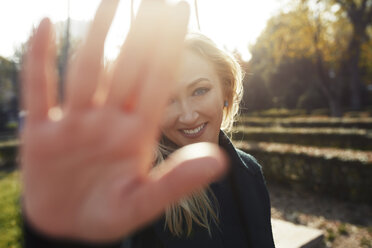 Porträt einer lächelnden blonden Frau, die ihre Hand in einem Park hebt - AZF00103