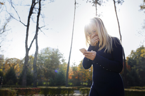 Lachende blonde Frau, die in einem herbstlichen Park auf ihr Smartphone schaut, lizenzfreies Stockfoto