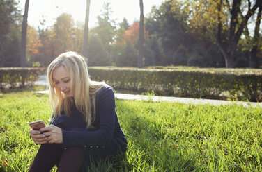 Lächelnde blonde Frau sitzt auf einer Wiese im herbstlichen Stadtpark und benutzt ihr Smartphone - AZF00106