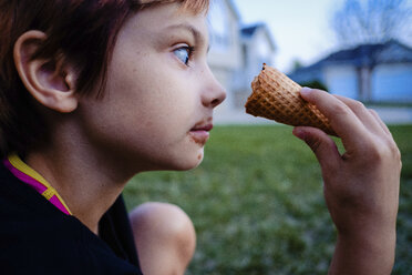 Side view of girl looking at ice cream cone while sitting in yard - CAVF49167