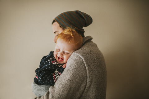 Seitenansicht eines Vaters, der seinen fröhlichen Sohn trägt, während er zu Hause an der Wand steht, lizenzfreies Stockfoto