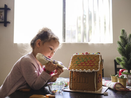 Seitenansicht eines Mädchens, das ein Lebkuchenhaus auf einem Tisch zu Hause glasiert - CAVF49219