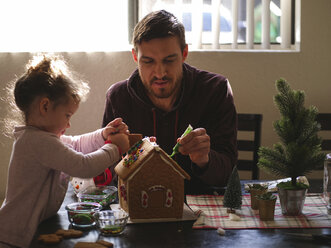 Daughter with father decorating gingerbread house on table at home - CAVF49220