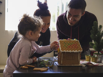 Happy parents looking at daughter decorating gingerbread house on table - CAVF49222