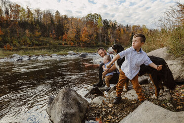 Vater mit Söhnen und Hund genießt am Fluss gegen bewölkten Himmel im Wald - CAVF49224