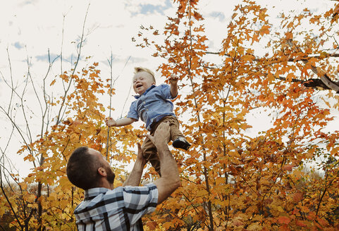 Low angle view of father throwing son while standing against sky in forest during autumn - CAVF49228