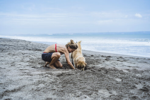 Full length of woman and Labrador Retriever digging on shore at beach - CAVF49238