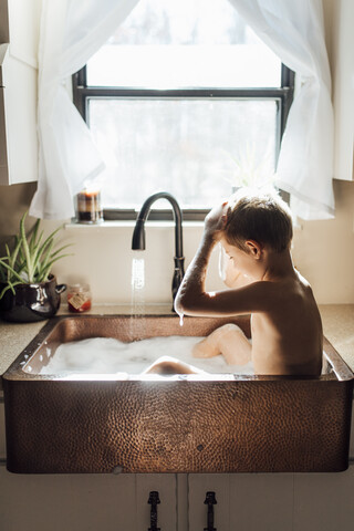 Shirtless boy taking bath while sitting in kitchen sink at home stock photo