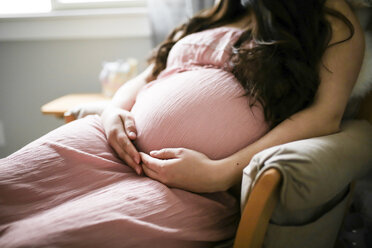 Midsection der schwangeren Frau berühren Bauch beim Sitzen auf Stuhl zu Hause - CAVF49264