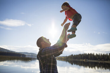 Seitenansicht eines spielerischen Vaters, der seinen Sohn in die Luft wirft, gegen einen See und Himmel an einem sonnigen Tag - CAVF49280