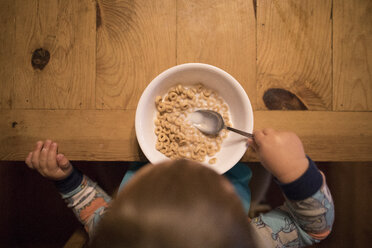 Draufsicht auf einen kleinen Jungen, der einen Löffel in einer Frühstücksschüssel auf einem Holztisch zu Hause hält - CAVF49288