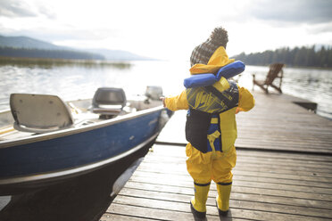 Rückansicht eines kleinen Jungen, der einen Regenmantel und eine Schwimmweste trägt, während er auf einem Holzsteg über einem See steht - CAVF49290