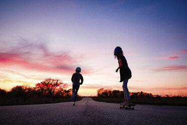 Silhouette girl skateboarding while sister riding push scooter on road against sky during sunset - CAVF49295