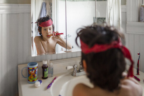 Hemdloser Junge mit rotem Stoff auf dem Kopf, der sich die Zähne putzt, während er auf sein Spiegelbild schaut - CAVF49319