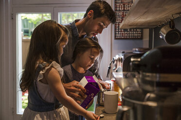 Töchter stehen mit Vater, der Milch in einen Becher am Küchentisch gießt - CAVF49327