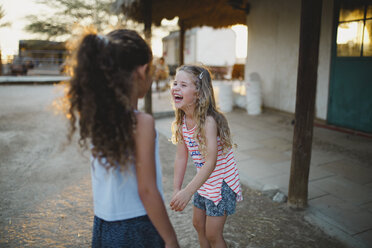 Mädchen lacht, während sie mit ihrer Schwester auf einem Bauernhof bei Sonnenuntergang steht - CAVF49364