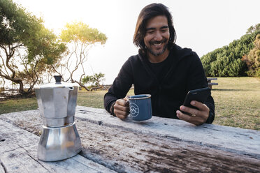 Glücklicher Mann mit Kaffee und Mobiltelefon auf einem Holztisch, während er gegen den Himmel sitzt - CAVF49369