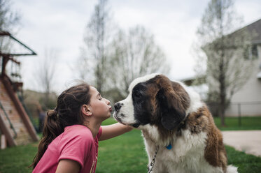 Glückliches Mädchen, das seinen Hund küsst, während es auf einer Wiese gegen den Himmel im Hof sitzt - CAVF49395