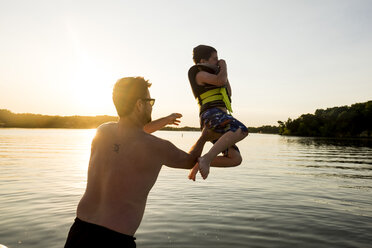 Rear view of shirtless father throwing son in lake against clear sky during sunset - CAVF49399