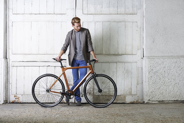 Full length of young man holding bicycle while standing against closed white doors on footpath - CAVF49408